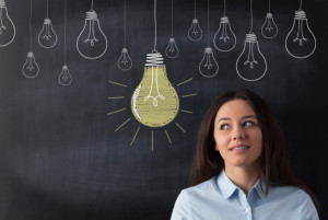 Young business woman standing with her back against a blackboard. She is looking to a big glowing light bulb.